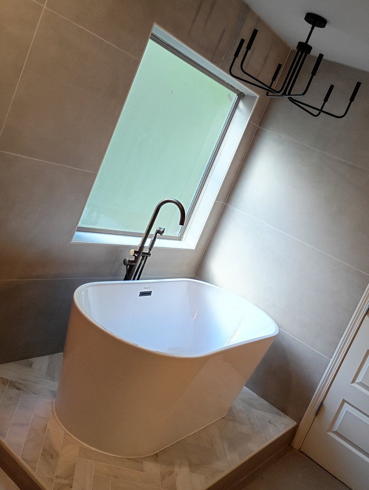 Finished bathroom with a freestanding tub set on a herringbone marble platform, large-format wall tile and a matte black chandelier.
