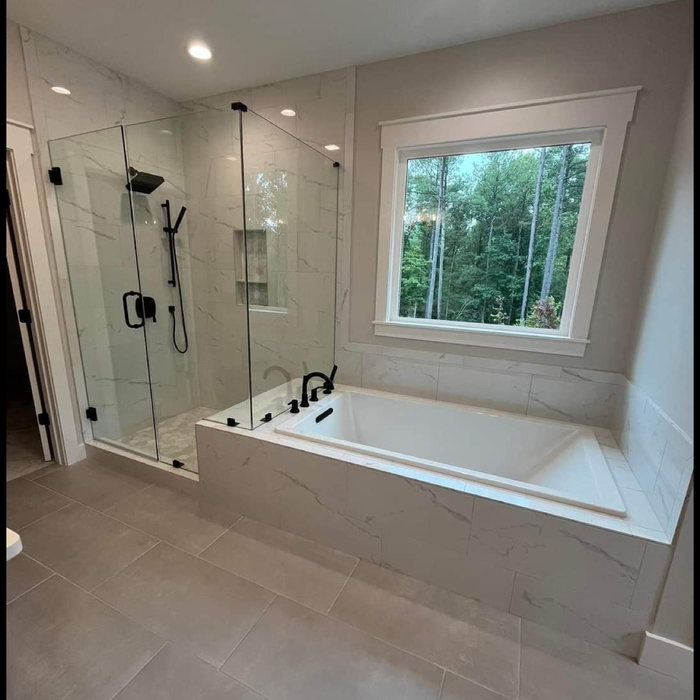 Primary bathroom with a frameless glass shower, marble-look tile and a drop-in tub set beneath a window looking into pine woods.