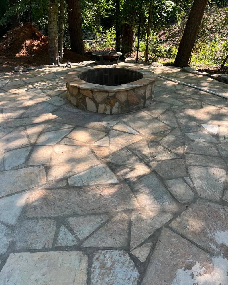 Irregular flagstone patio laid around a circular stone fire pit under mature trees.