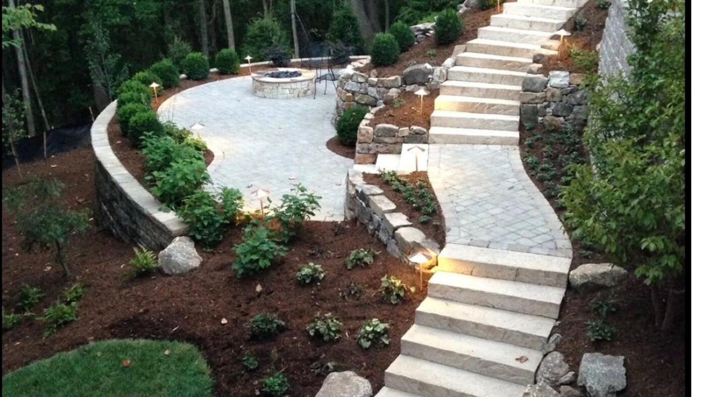 Curved paver terrace with a stone fire pit, granite stair treads and landscape lighting set into a wooded slope at dusk.