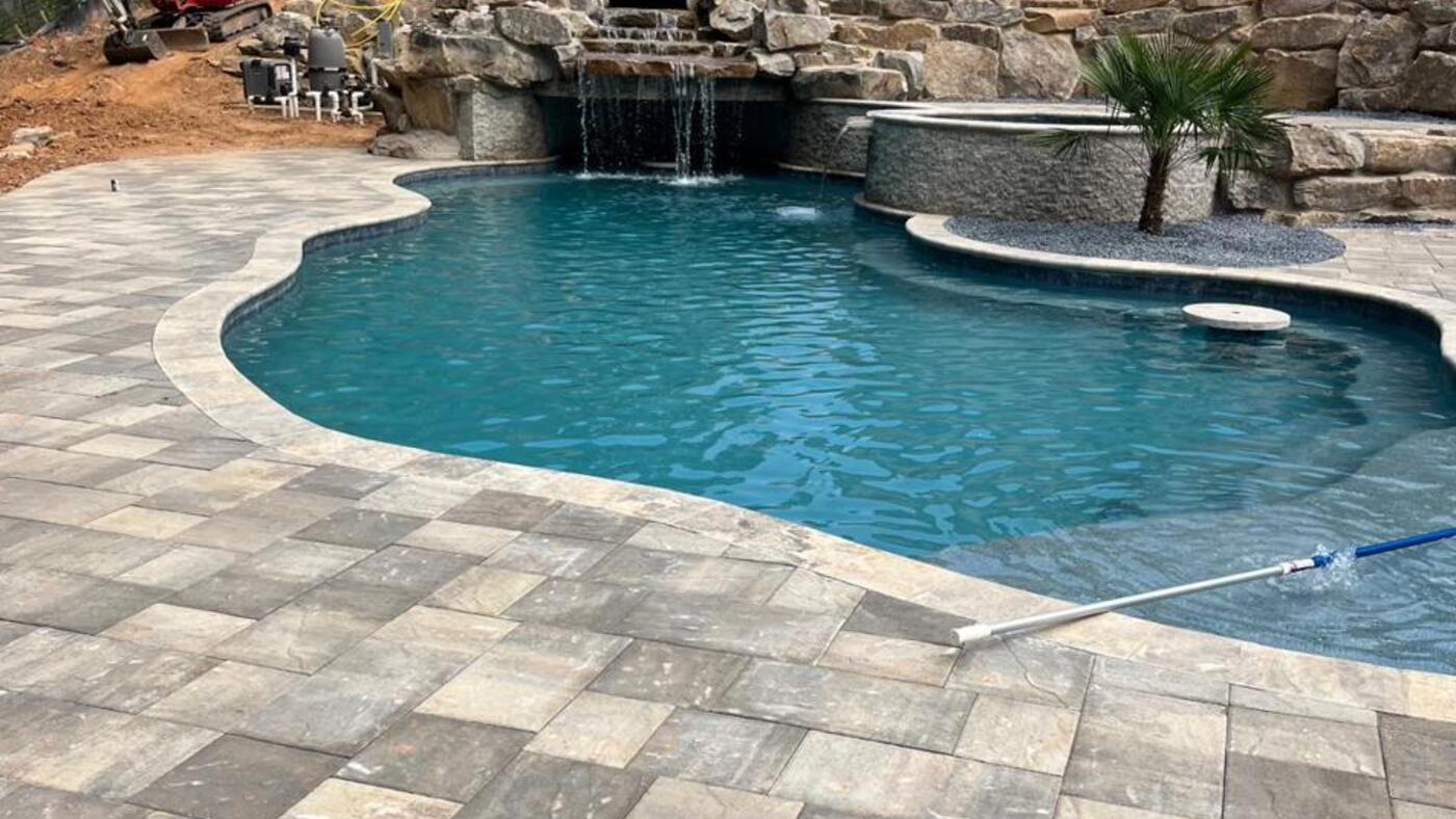 Freeform swimming pool with a stone waterfall feature, edged by a large-format travertine paver deck.