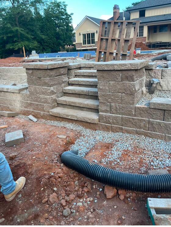 Corrugated drainage pipe run behind a block retaining wall on a gravel bed during backfill.