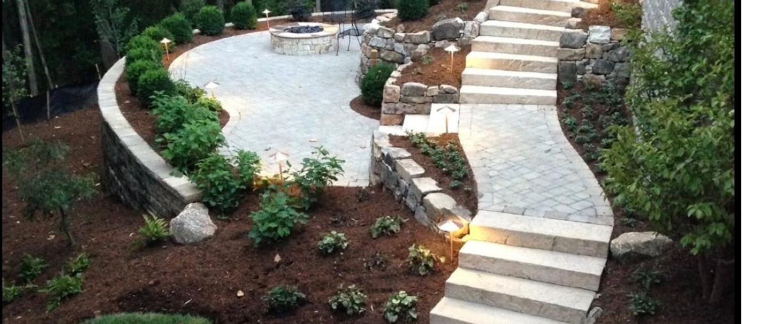Wooded backyard transformed with a curved paver terrace, stone fire pit, granite stair treads climbing the slope, boulder-set retaining walls, fresh planting beds and low landscape lighting.