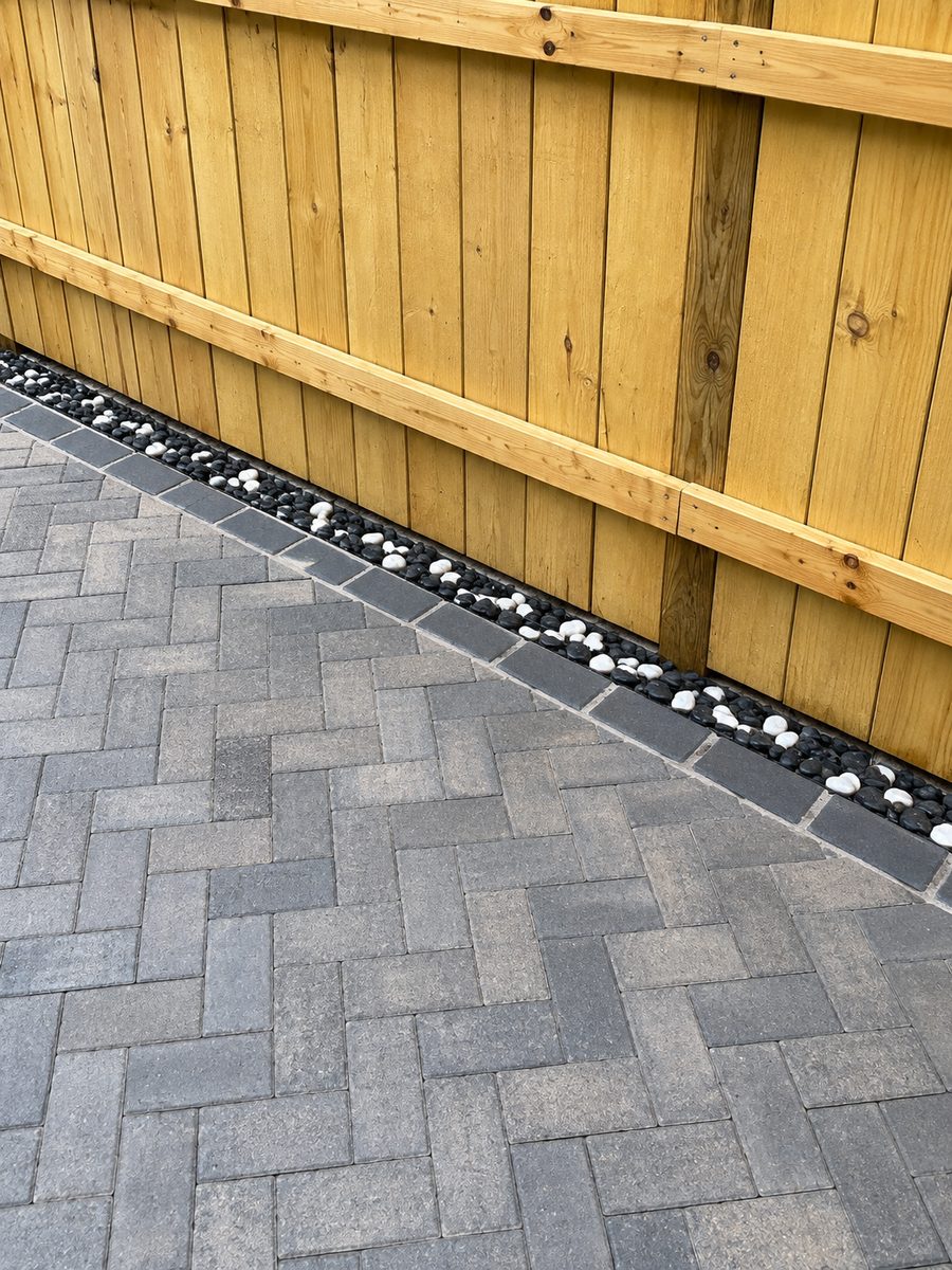 Finished grey herringbone paver patio with a charcoal border course and a black-and-white decorative stone reveal along a wood privacy fence.