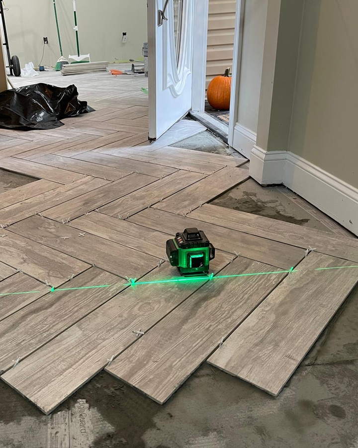 Wood-look tile dry-laid in a herringbone pattern across an entry hall, with a green laser level projecting alignment lines toward an open front door.